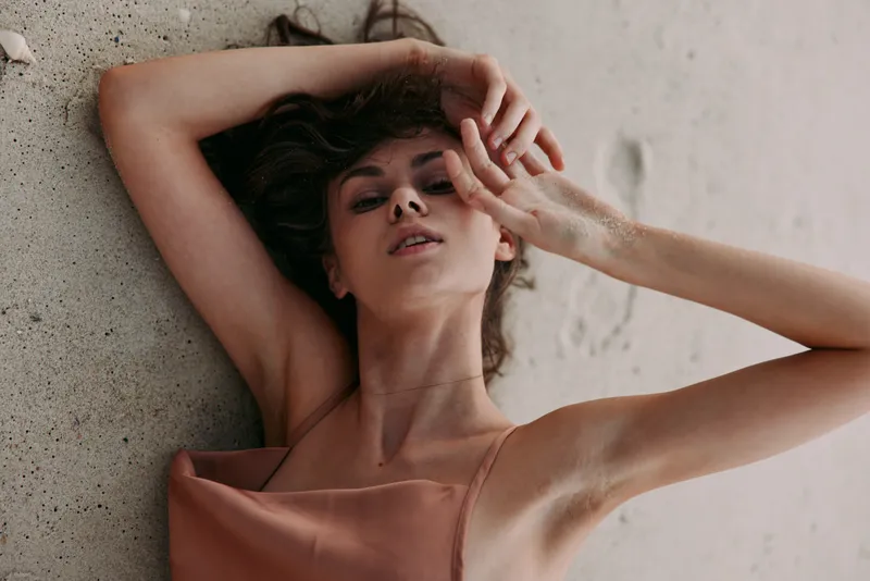 Portrait of a relaxed woman leaning against a textured wall, soft light highlights her serene expression, muted satin top, and casual pose creating an intimate artistic mood.