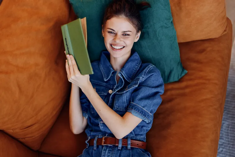 Молодая женщина в джинсовой рубашке читает книгу, сидя на диване дома, наслаждаясь досугом в уютной и комфортной гостиной, вид сверху