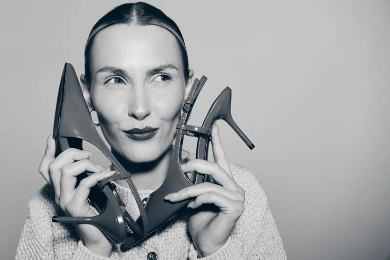 Young white woman with fair skin holds high heel shoes near face, smiling with playful expression against grey background in studio lighting.