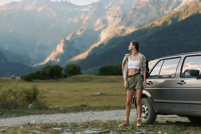 A woman in casual summer clothes stands beside an off-road vehicle in a mountainous landscape, enjoying the outdoor lifestyle and nature adventure concept.