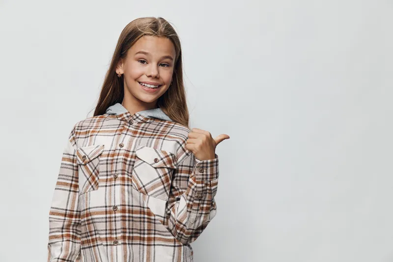Teenage girl wearing plaid shirt smiling and pointing thumb back isolated on white background, happy expression with casual style and confident pose in studio.