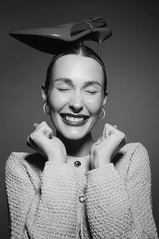 Joyful Caucasian woman with fair skin smiles with closed eyes wearing a high heel shoe on her head and cozy knitted sweater in studio lighting.