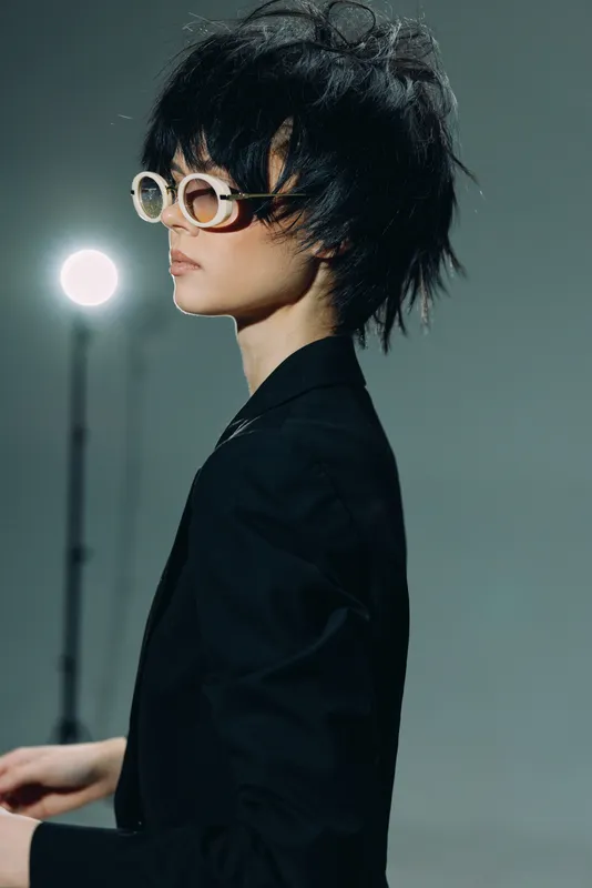 Stylish woman wearing retro sunglasses and black jacket with dark messy hair looking sideways in studio light.