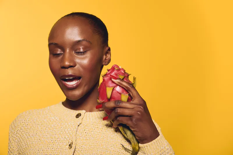 African woman with dark skin wearing yellow sweater holds dragon fruit near face with eyes closed on bright yellow background. Concept of healthy lifestyle, exotic fruit, and natural beauty.