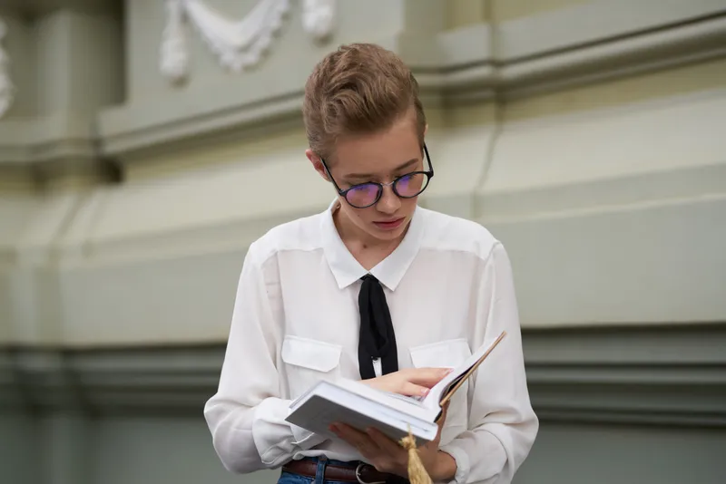 Счастливая студентка возле здания с книгой в руке и в очках на лице