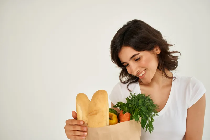 Жизнерадостная женщина в белой футболке держит пакет с продуктами из супермаркета