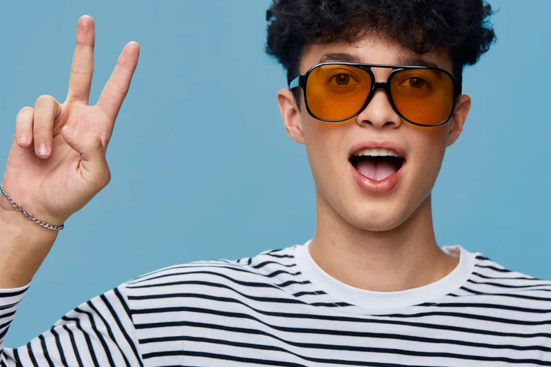 Young man showing peace sign wearing orange glasses and striped shirt against blue background, expressing happiness and casual style with curly hair.
