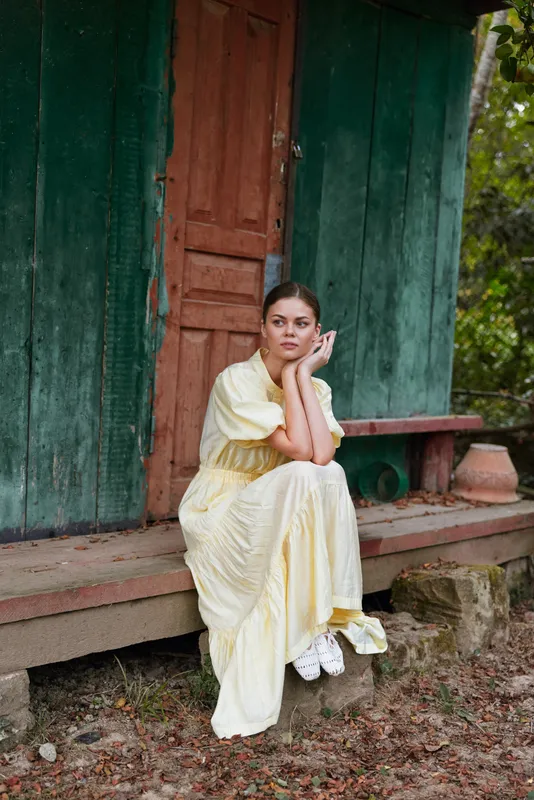 Woman in a pale yellow dress sits on wooden steps outside a rustic building, thoughtful and serene, soft natural light highlighting a calm, introspective mood