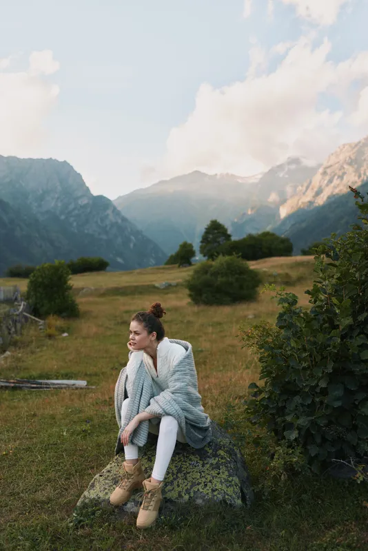 Young woman sits on a rock in a mountain meadow, wearing a light cardigan and beige boots, enjoying tranquil nature and fresh alpine air, her relaxed pose conveying calm and contemplative mood amid