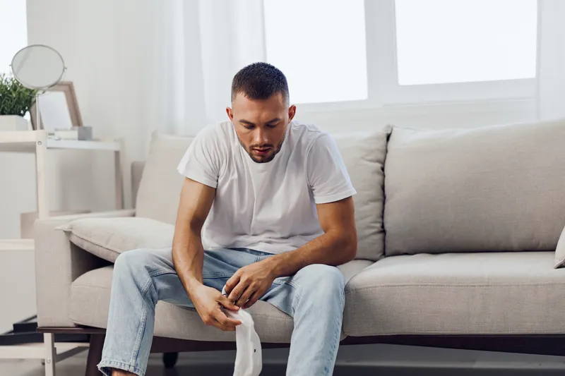 Pensive man sitting on a couch, contemplating life decisions while holding a piece of clothing. The modern interior offers a calm and reflective ambiance.