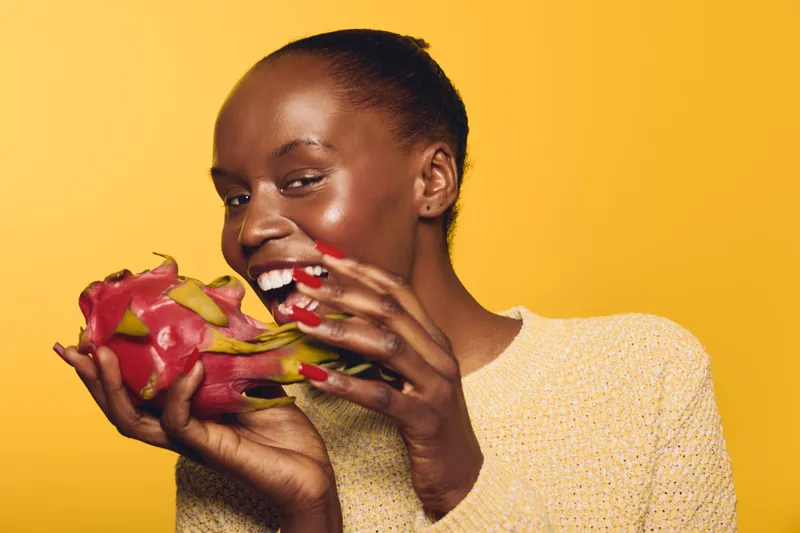 Smiling African woman with dark skin holds a dragon fruit near her face on yellow background. Concept of healthy eating, exotic fruit, natural beauty, and joyful lifestyle.