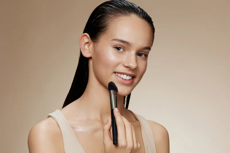 Young woman applying makeup with a brush, displaying a natural look and a radiant smile against a soft neutral background, highlighting beauty and skincare concepts