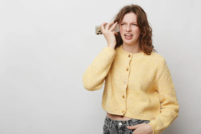 Angry young woman in yellow sweater holds smartphone to ear, expressing frustration and annoyance. Isolated on white background. Concept of communication problems and emotional stress.