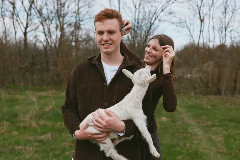 Cheerful couple outdoors with a white goat kid, sharing a playful moment in a sunlit meadow, warm breeze, smiling faces, tender affection, vibrant natural colors, rustic park backdrop