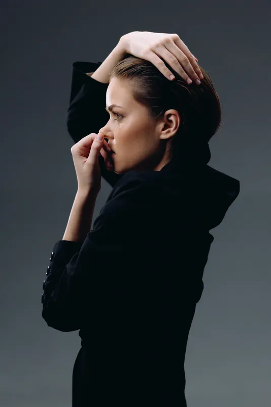 Serious woman in black sweater posing with hand on head against dark background indoors.