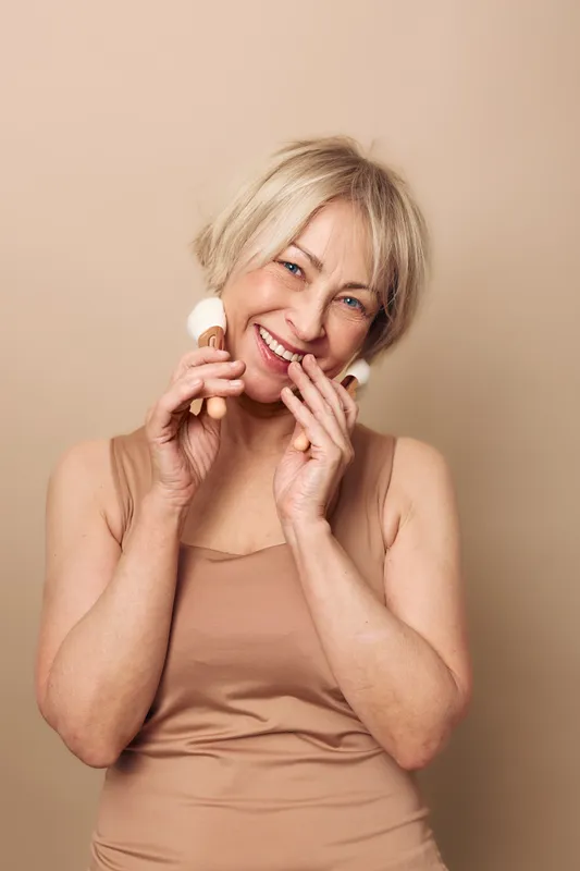 Smiling mature woman with light hair uses facial brushes on beige background. Concept of skincare, natural beauty, self-care, and wellness for aging skin with joyful expression.