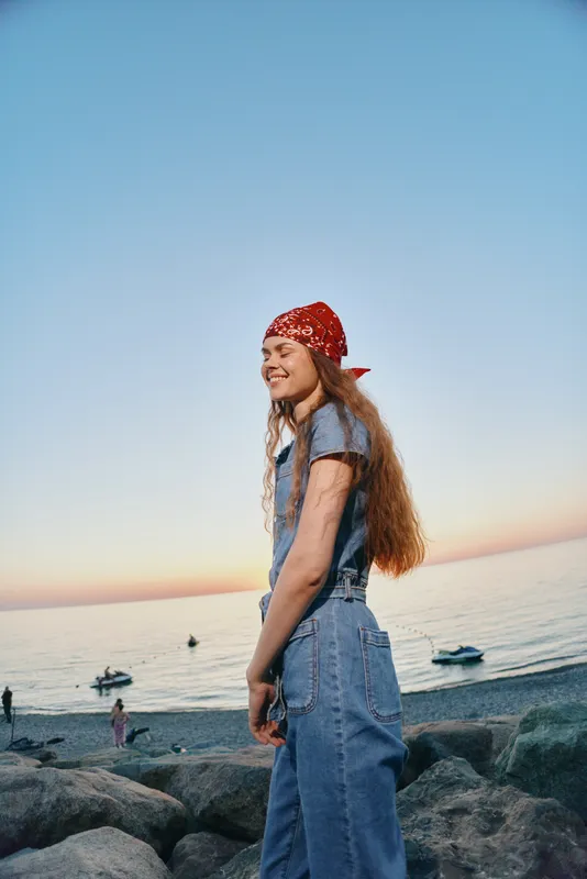 smiling woman in denim outfit with red bandana enjoying sunset by the seaside with rocky shore and calm ocean background