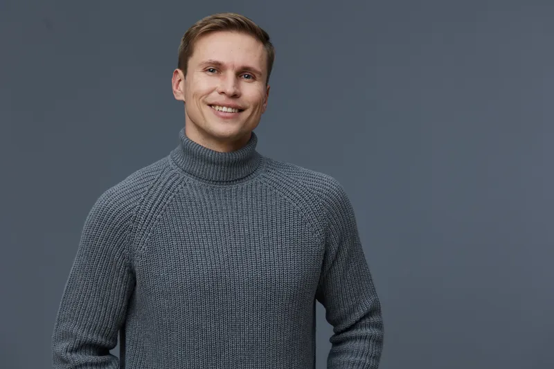 Smiling man with light skin, short brown hair, wearing gray turtleneck sweater looking at camera isolated on plain blue background studio portrait. People lifestyle concept