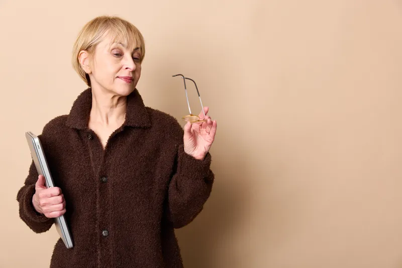 Elderly woman, beige studio background, smiling and holding a notebook and glasses, showing thinking for education and communication, calm indoor portrait with copyspace