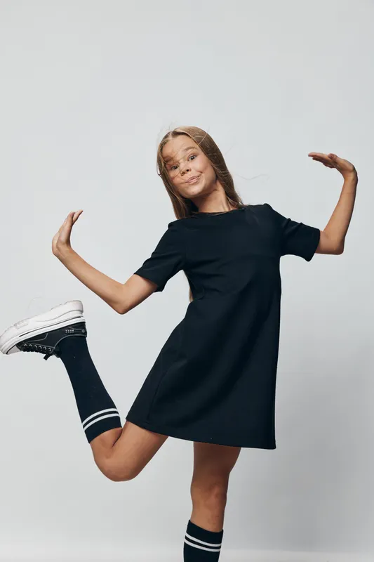 Girl in black dress and sneakers posing with playful expression on gray background for youth fashion and lifestyle portrait. Casual outfit with knee socks and fun pose for studio photo.