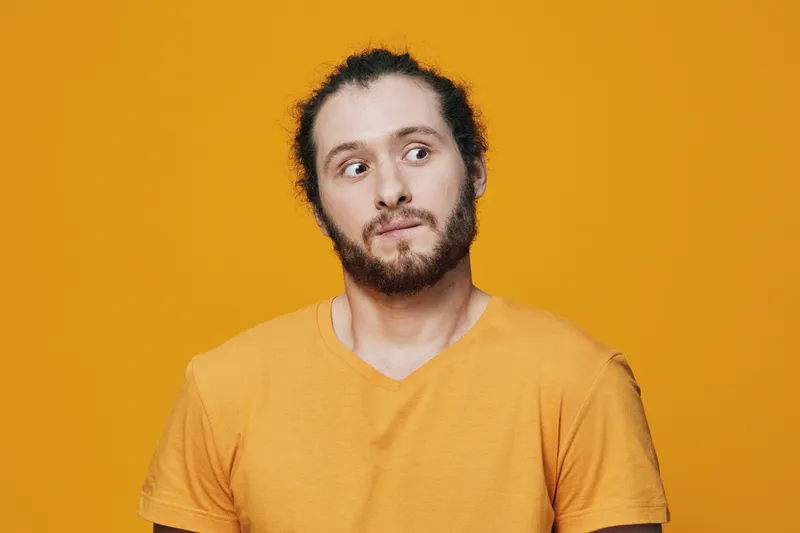 A young man with a beard in a yellow t shirt showing a perplexed expression against a vibrant yellow background, embodying curiosity and surprise in a modern setting