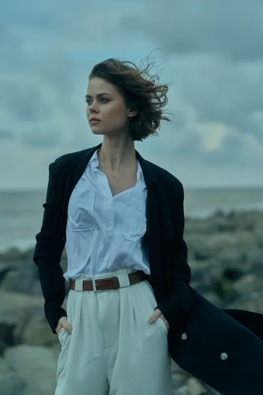 Woman in blazer, white shirt and pants stands on a rocky coastline, wind in hair, looking to the horizon, showcasing chic outdoor fashion and coastal mood
