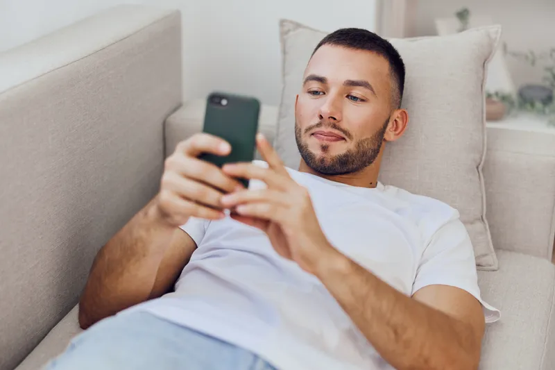Relaxed man using smartphone while lounging on a cozy couch, enjoying modern technology in a stylish living room. Perfect blend of comfort and casual lifestyle.