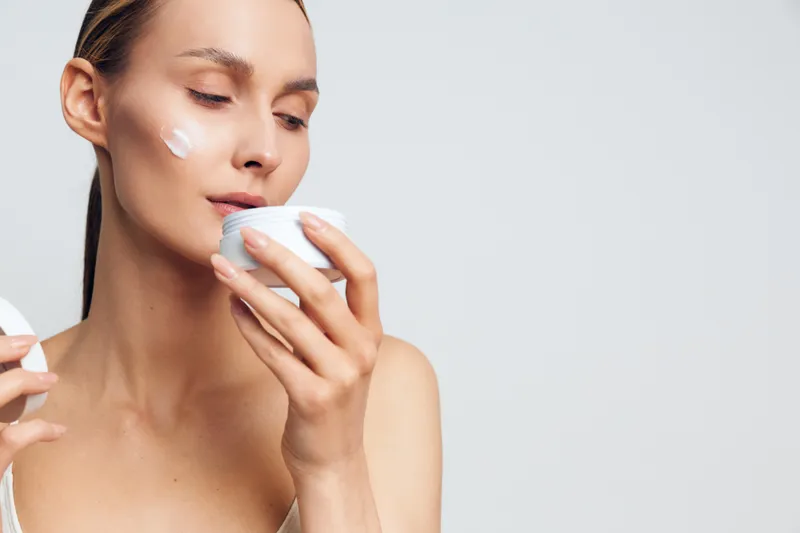 A young woman with clear skin applies cream on her cheek while smelling a white jar of skincare product in a bright studio. Concept for beauty, self care, and skincare routine.