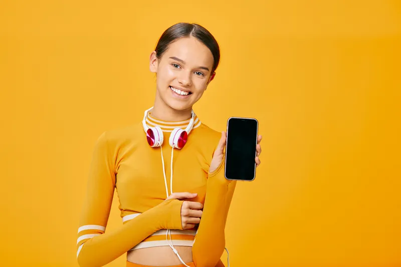 Fitness minded woman in a bright yellow outfit stands against a sunny backdrop, holding a smartphone and wearing headphones, ready for a quick workout, dance or training session, showcasing casual