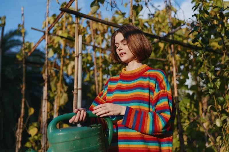 Woman in a colorful striped sweater holds a green watering can in a backyard garden, young adult with bob haircut smiling softly, casual outfit, outdoor lifestyle and gardening.