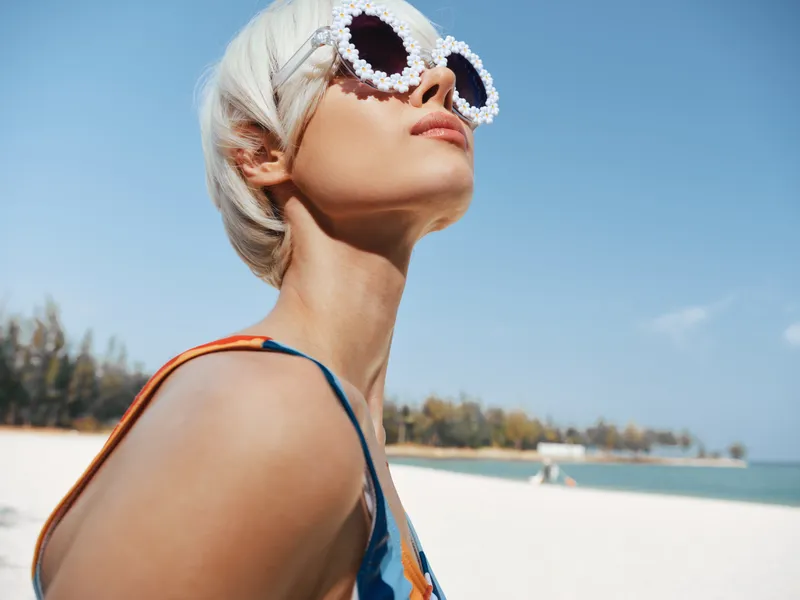 Close up of a stylish woman on a bright beach, wearing oversized shell framed sunglasses and a colorful swimsuit, sunlit skin and a clear blue sky above, emphasizing summer fashion and confident pose.