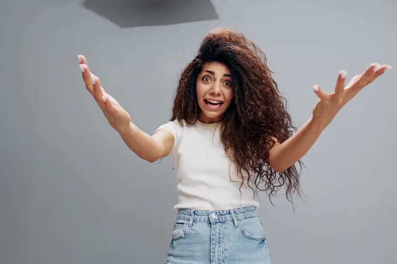 Крупный план удивленной потрясающей молодой женщины, которая с вытаращенными глазами замечает что-то ужасное впереди, разводит руки в стороны перед камерой, позирует изолированно на сером фоне