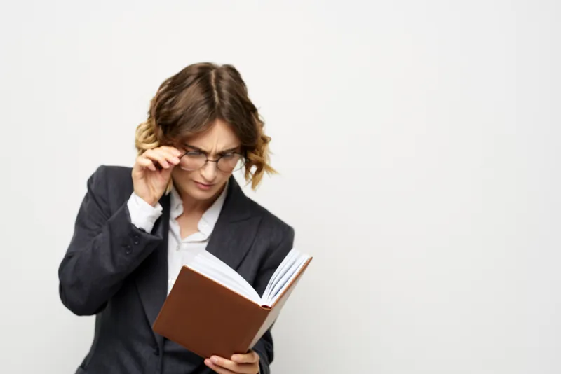 Женщина за работой с книгой в руке на светлом фоне, в классическом костюме и очках
