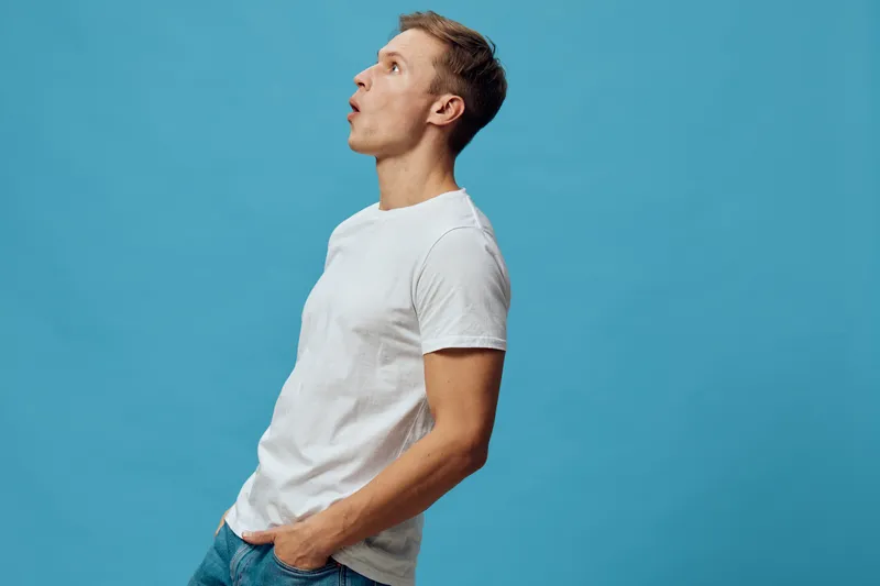 Casual man in white t-shirt looking up against vibrant blue background, relaxed pose, side view, modern lifestyle, minimalistic style, male portrait, studio shot, white wall, youthful appearance.