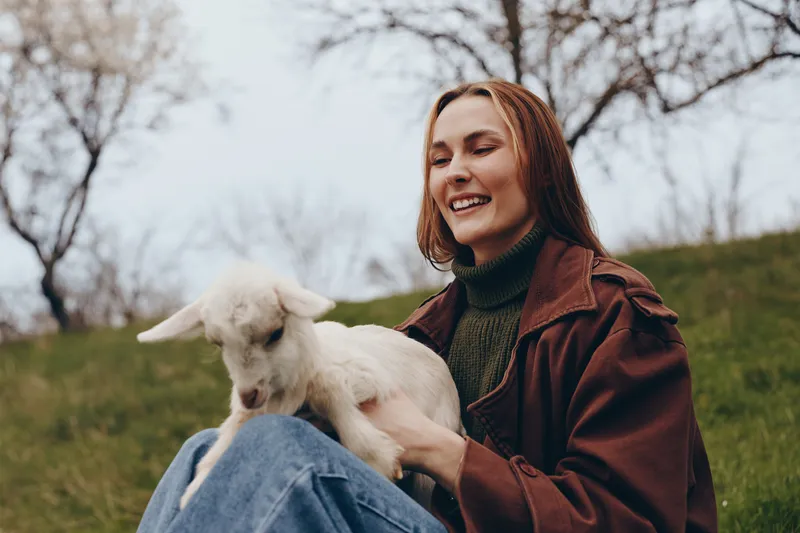 Joyful woman outdoors, caressing a playful goat kid in a sunlit field, relaxed jacket sweater attire, soft breeze, earthy colors, pastoral nature scene