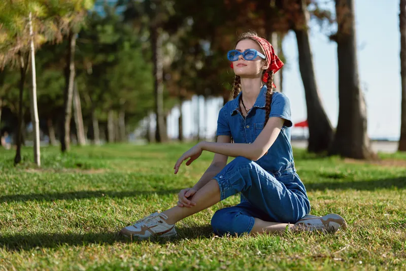 young woman sitting on grass wearing casual denim outfit and sunglasses enjoying sunny day in outdoor park with trees and green lawn background