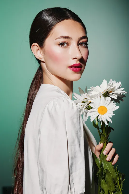 Young woman with long dark hair in white blouse holds white daisies against green background. Glowing skin and vibrant red lips accentuate natural beauty and elegant fashion style.