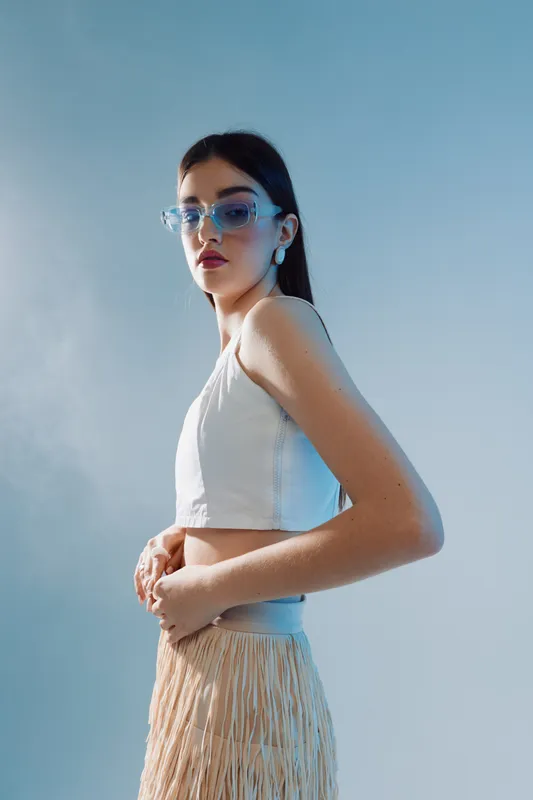 Stylish woman in a white crop top and beige skirt poses confidently wearing blue glasses. Studio shot on a light blue background. Concept of woman beauty fashion and elegance.