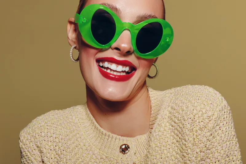 Smiling woman with bright red lipstick wears large green sunglasses and beige knitted sweater. Close-up portrait on neutral background. Concept of fashion, style, and joyful mood.