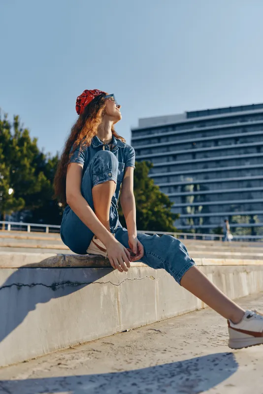Lifestyle woman in modern boho-western denim street style sitting outdoors in film color, relaxed pose, sunny day, urban background, casual fashion look