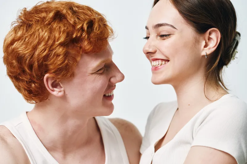 Young couple with red hair and brunette smiling at each other in closeup portrait. Positive emotions, friendship and happiness captured in casual white clothing on white background.