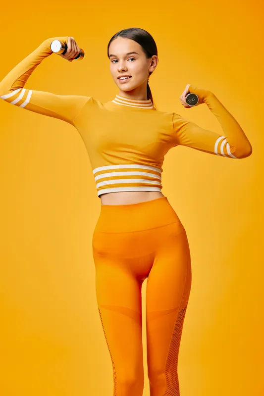 Woman fitness athlete in orange sportswear lifts dumbbells against a vibrant orange backdrop, showcasing strength, balance, and dynamic movement with confident posture and focused expression.