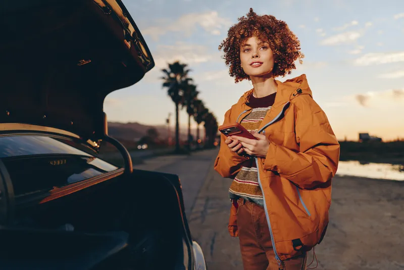 Woman lifestyle outdoors at sunset beside a parked car trunk open, curly haired traveler in orange jacket with backpack holds phone, ready for travel departure on road