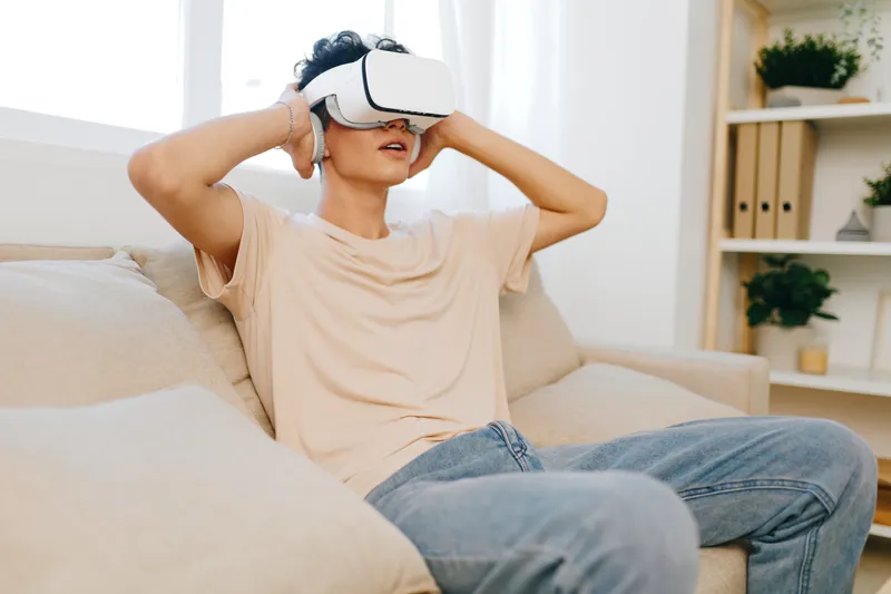 Сцена игры в виртуальной реальности: молодой человек в шлеме VR удобно сидит на бежевом диване
