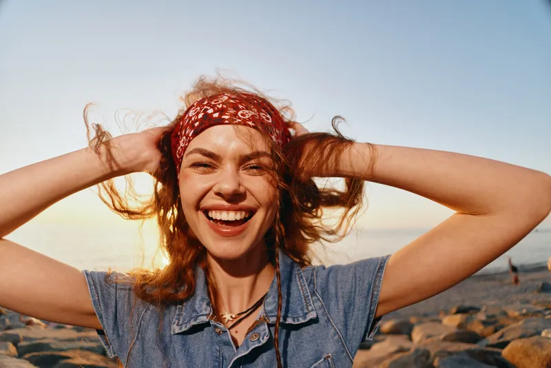 smiling woman wearing red headband and denim shirt enjoying sunny beach with ocean background, long wavy hair, natural light, cheerful mood outdoors