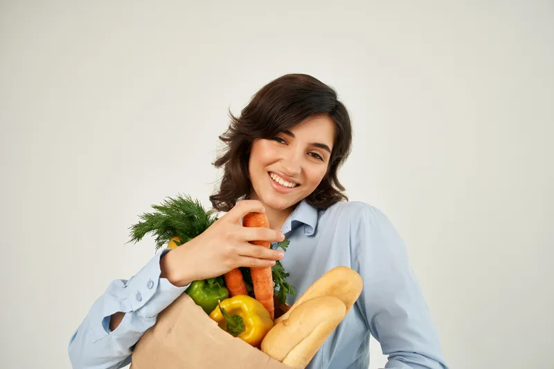 Жизнерадостная женщина с пакетом продуктов, включая овощи, на фоне доставки из супермаркета
