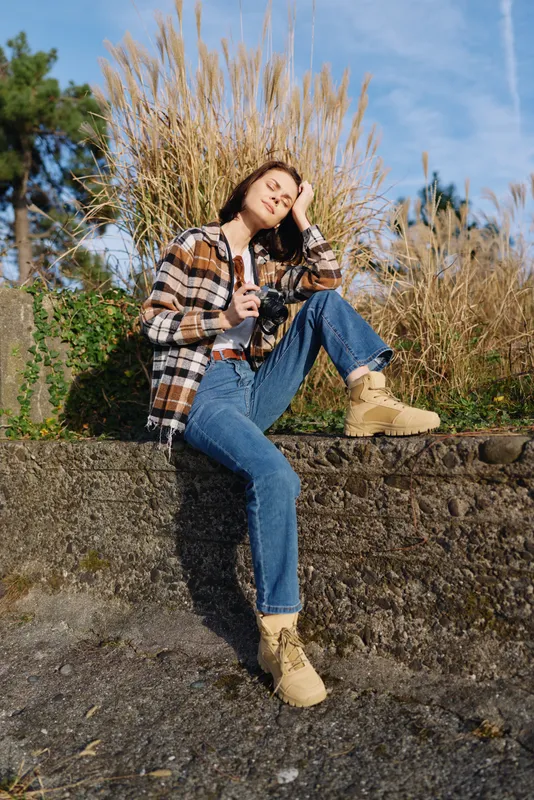 Outdoor woman posing in casual fashion on a stone wall, plaid jacket, jeans and boots under a sunny sky, embracing a relaxed lifestyle, nature backdrop and candid mood.