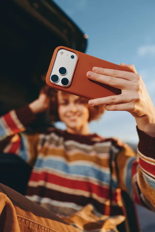 Woman holding smartphone for a selfie, close hand view with brown case, family moment with a smiling child behind. Warm street atmosphere, lifestyle travel feeling, candid portrait