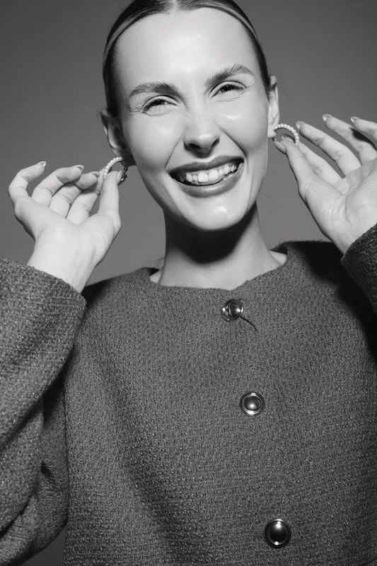 Happy white woman with light skin smiles broadly, holding earrings near her face in studio. Concept of joy, fashion, and beauty in black and white photography.