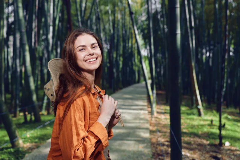 Woman smile with backpack on forest path, happy traveler enjoying nature and trees. Portrait of young woman in orange shirt hiking on peaceful trail with joyful expression and sunlight.